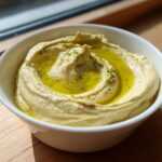 A close-up of creamy, light yellow hummus in a white bowl, topped with a swirl of olive oil and sprinkled herbs.