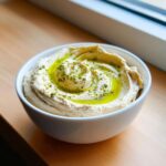 A close-up of creamy homemade hummus swirled in a white bowl, topped with olive oil and green spices.