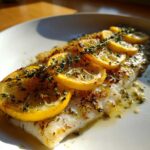 Close-up of a flaky Baked Tilapia fillet topped with overlapping lemon slices and fresh thyme sprigs.