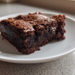 A close-up view of a rich, dark, fudgy brownies slice with a shiny, cracked top, served on a white plate.
