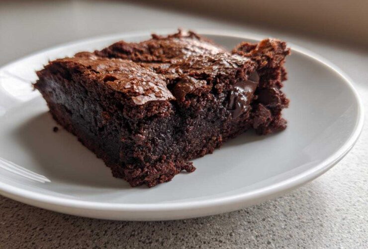 A close-up view of a rich, dark, fudgy brownies slice with a shiny, cracked top, served on a white plate.