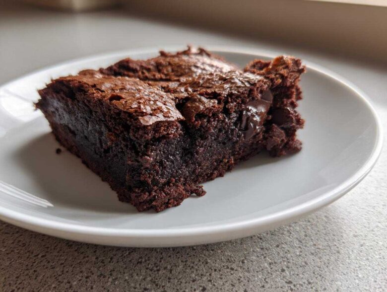 A close-up view of a rich, dark, fudgy brownies slice with a shiny, cracked top, served on a white plate.