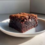 A close-up of a rich, dark, fudgy brownies square with visible chocolate chips on a small white plate.