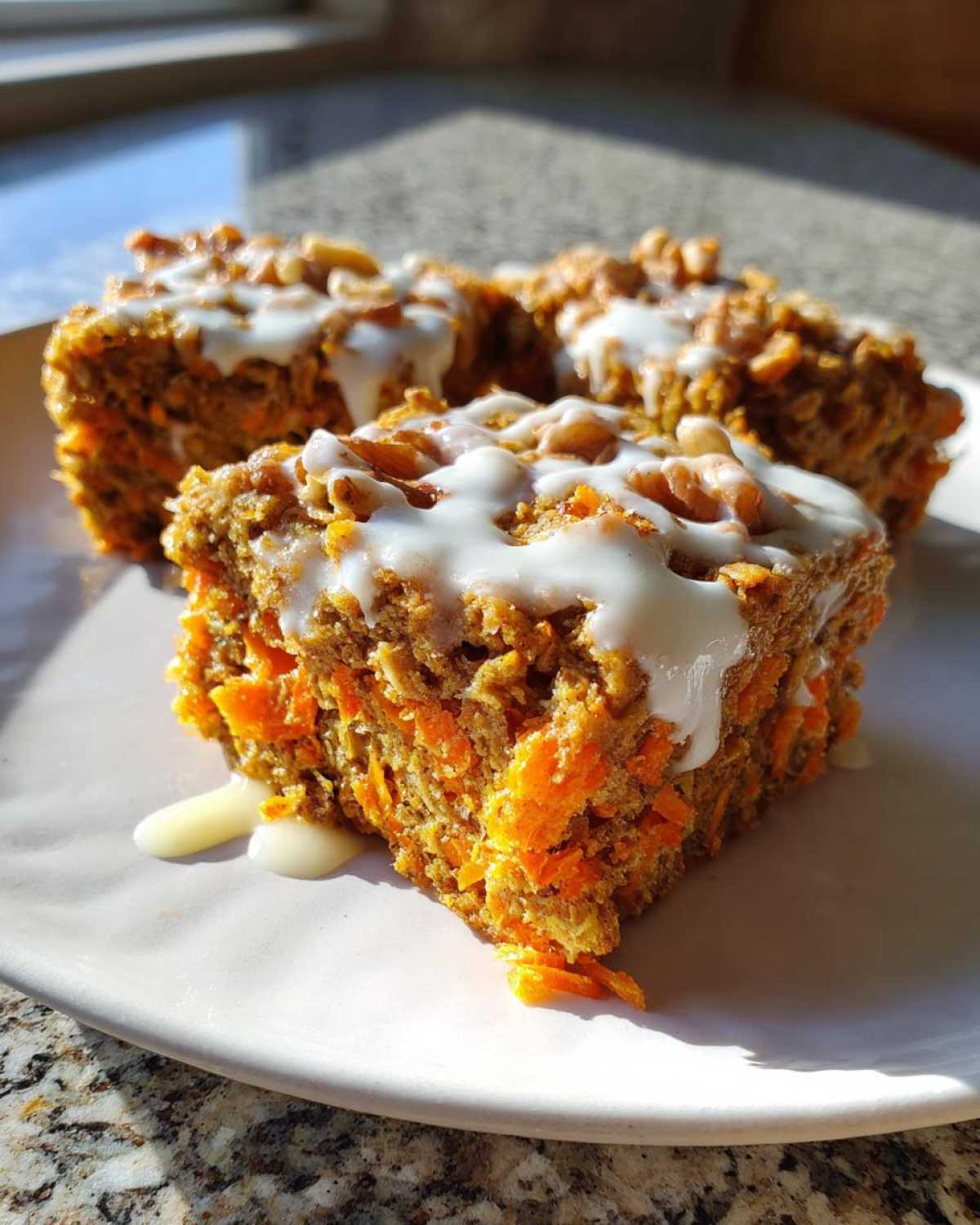 Three squares of Gluten-Free Carrot Cake Baked Oats topped with white icing and nuts on a white plate.