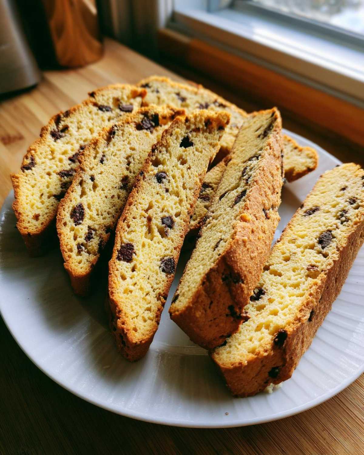 Several golden-brown slices of Gluten-Free Chocolate Chip Biscotti arranged on a white plate near a window.