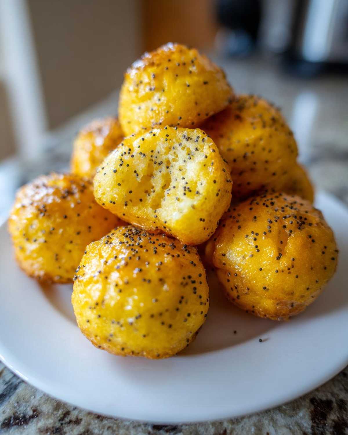 A stack of golden, glazed Gluten-Free Lemon Poppy Seed Donut Holes sprinkled with poppy seeds on a white plate.