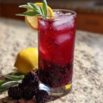 A tall glass of vibrant red Iced Blackberry Sage Tea filled with ice, garnished with lemon and sage.