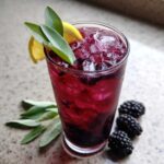 A tall glass filled with vibrant Iced Blackberry Sage Tea, ice, and garnished with fresh sage leaves and a lemon slice.