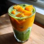 A tall glass showing the vibrant layers of an Iced Mango Coconut Matcha drink with mango chunks on top.