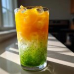 A tall glass showing distinct layers of bright green matcha, white coconut, and vibrant orange mango in an Iced Mango Coconut Matcha.