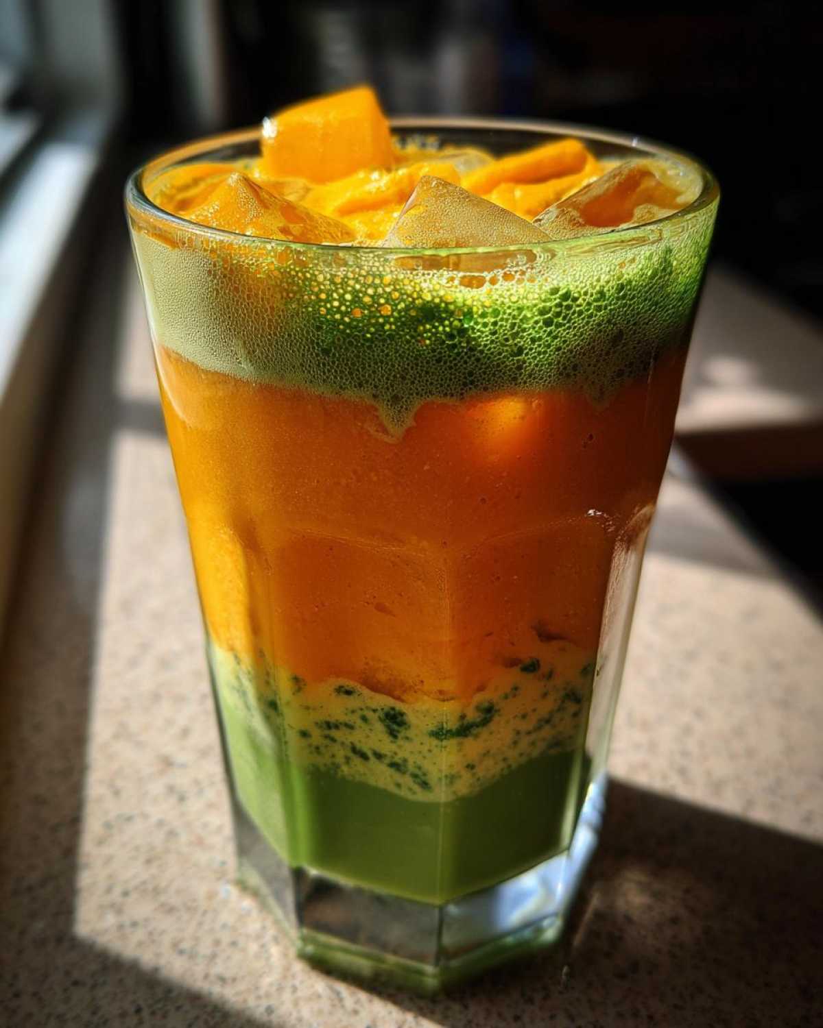 Close-up of a tall glass showing distinct layers of green matcha, orange mango, and foam in an Iced Mango Coconut Matcha.