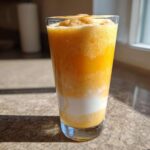 A tall glass filled with a layered Iced Orange Creamsicle Smoothie, showing bright orange frozen layers and a white creamy layer.