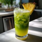 A tall glass filled with a vibrant green Iced Pineapple Matcha Spritz, ice, and garnished with a fresh pineapple wedge.