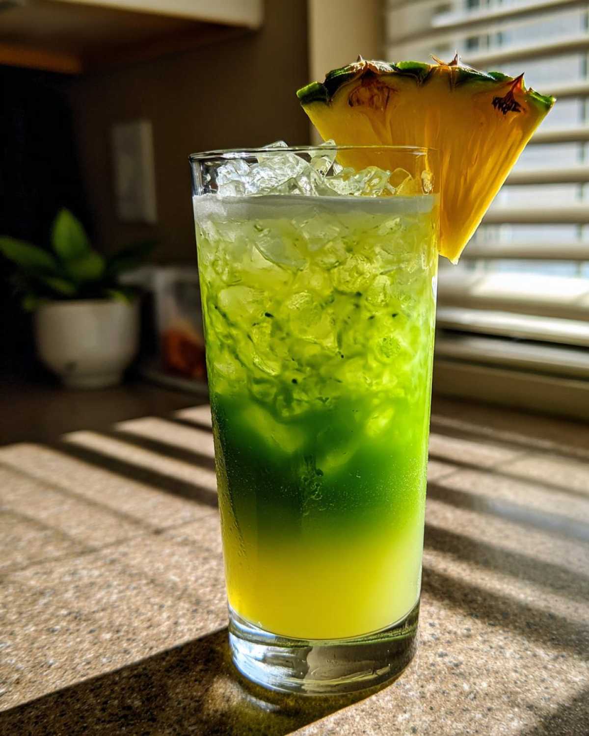 A tall glass filled with a layered Iced Pineapple Matcha Spritz, featuring yellow pineapple juice and green matcha, garnished with a pineapple wedge.