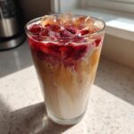 Close-up of a tall glass filled with Iced Raspberry Vanilla Chai, showing layered milk, tea, and a bright raspberry topping over ice.