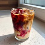 A tall glass of Iced Raspberry Vanilla Chai showing dramatic swirling layers of dark red raspberry syrup, creamy milk, and brown chai concentrate over ice.