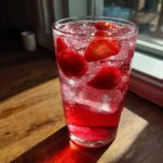 A tall glass filled with bubbly, vibrant Iced Strawberry Hibiscus Tea Lemonade, garnished with fresh strawberry slices and ice.