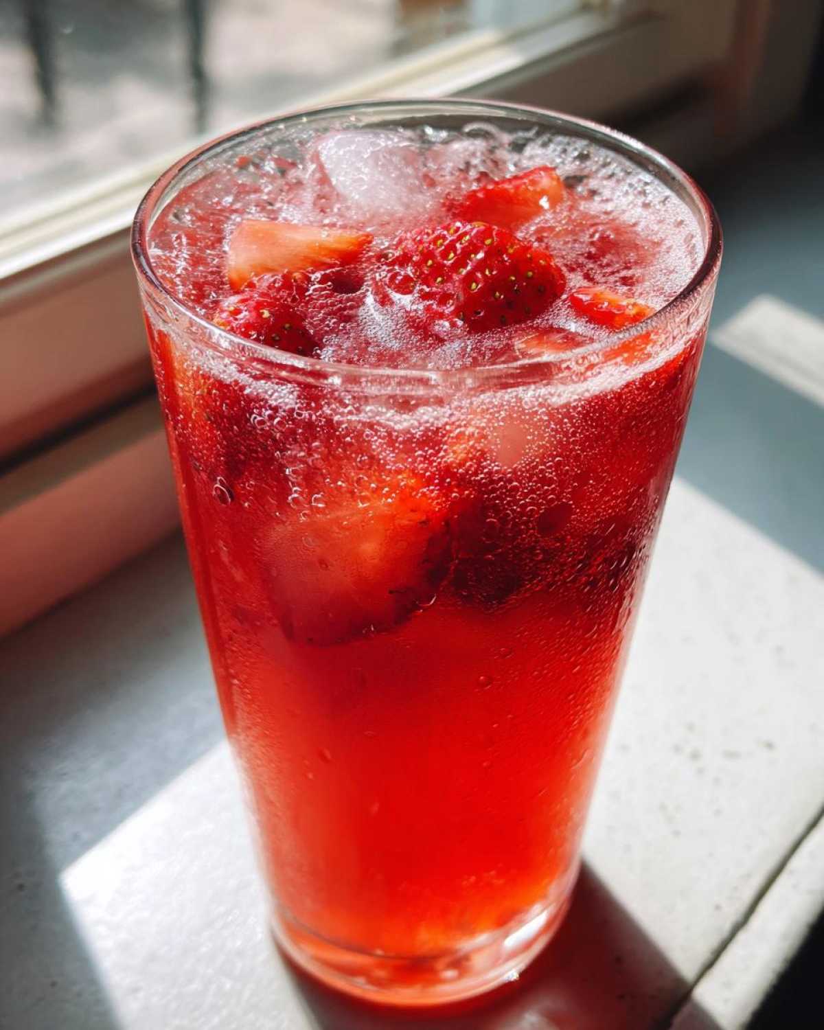 Close-up of a tall glass filled with vibrant red Iced Strawberry Hibiscus Tea Lemonade, ice, and fresh strawberry slices.