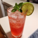 A tall glass filled with a bubbly, pink Iced Watermelon Lime Cooler, garnished with mint and a lime wheel.