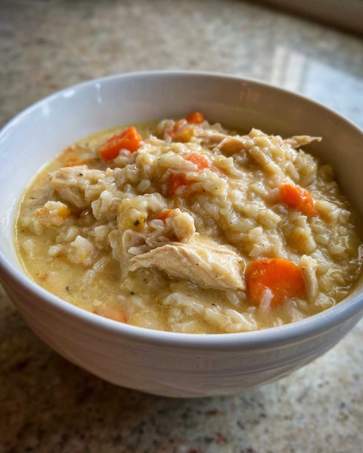 Close-up of a white bowl filled with creamy Instant Pot Creamy Chicken and Rice Soup, featuring shredded chicken, rice, and bright orange carrots.