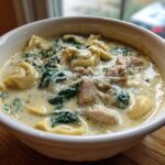 Close-up of a white bowl filled with Instant Pot Creamy Chicken Tortellini Soup, featuring tortellini, chicken chunks, spinach, and cheese.