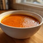 A close-up of a white bowl filled with bright orange Instant Pot Creamy Red Pepper Tomato Soup on a wooden counter.