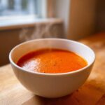 A steaming white bowl filled with vibrant orange Instant Pot Creamy Red Pepper Tomato Soup, seasoned with pepper.