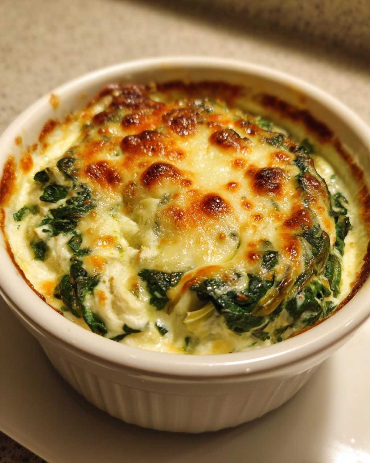 Close-up of a small ramekin filled with bubbling Instant Pot Creamy Spinach Artichoke Dip topped with melted, browned cheese.