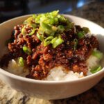 A close-up of a white bowl filled with white rice topped with saucy Instant Pot Korean Beef and garnished with sliced green onions.