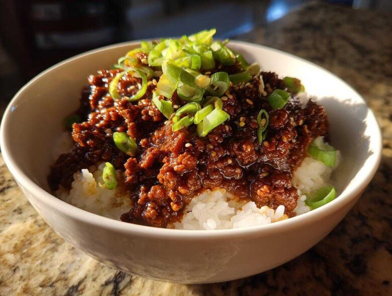 A close-up of a white bowl filled with white rice topped with saucy Instant Pot Korean Beef and garnished with sliced green onions.