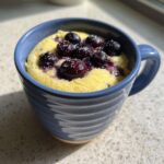 Close-up of a freshly made Keto Blueberry Lemon Mug Cake topped with fresh blueberries and sugar crystals in a blue mug.