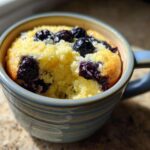 A close-up view of a fluffy Keto Blueberry Lemon Mug Cake baked in a blue ceramic mug, topped with dark blueberries.