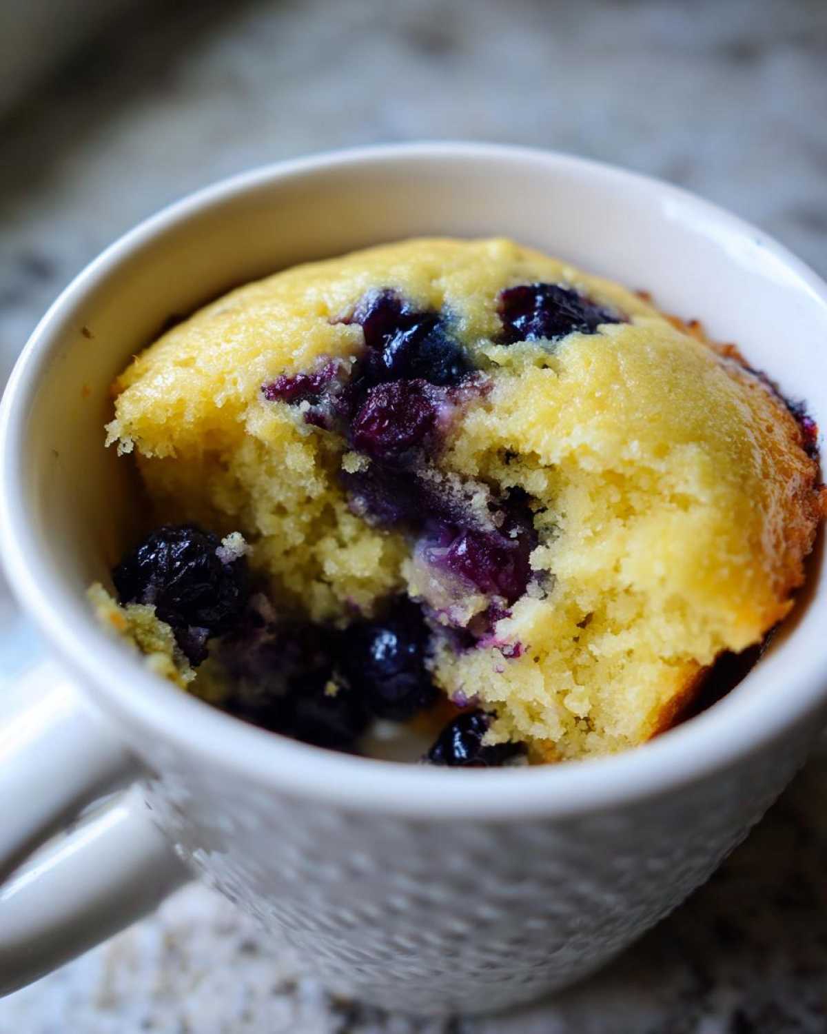 A close-up view of a fluffy Keto Blueberry Lemon Mug Cake with visible blueberries inside a white mug.