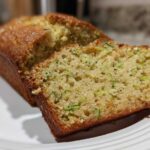 Close-up of a freshly baked Zucchini Bread loaf with one slice cut, showing moist texture and green shreds of zucchini.