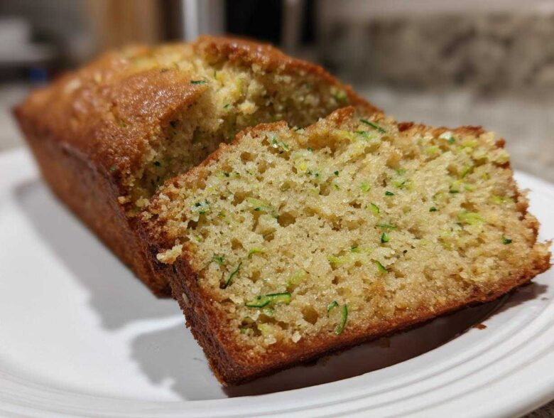 Close-up of a freshly baked Zucchini Bread loaf with one slice cut, showing moist texture and green shreds of zucchini.