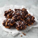 A pile of rich No-Bake Chocolate Covered Pretzel Clusters topped with flaky sea salt on parchment paper.