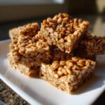 A stack of square No-Bake Peanut Butter Rice Cereal Bars glistening on a white plate.