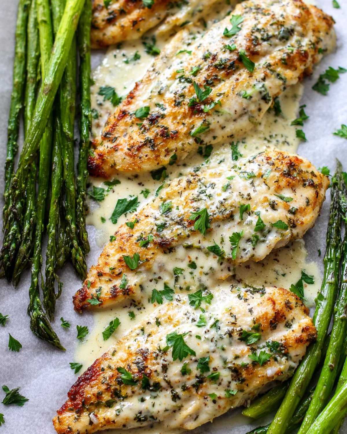 Three fillets of One-Pan Creamy Garlic Butter Tilapia covered in sauce and parsley, served alongside roasted asparagus.