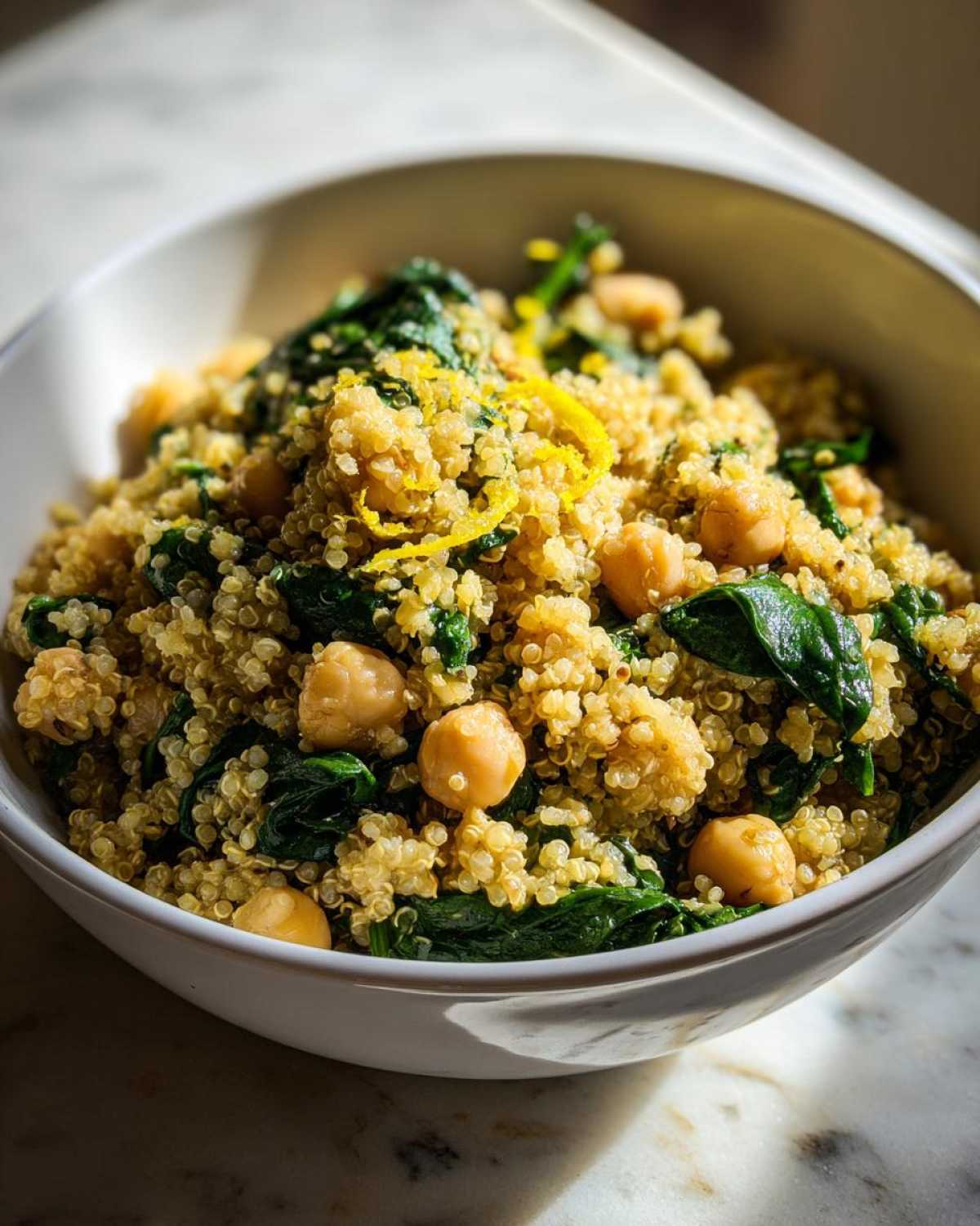 Close-up of a white bowl filled with the One Pot Chickpea Quinoa Recipe, mixed with spinach and topped with lemon zest.