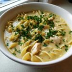 A close-up of a white bowl filled with One-Pot Creamy Lemon Chicken Noodle Soup, topped with parsley and lemon zest.