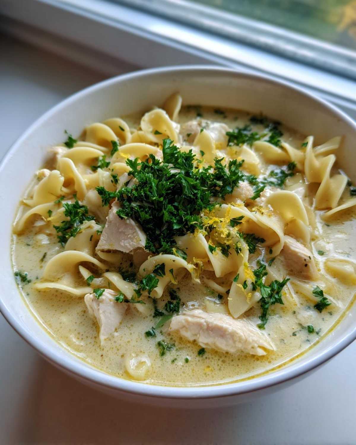 A close-up of a white bowl filled with One-Pot Creamy Lemon Chicken Noodle Soup, garnished with parsley and lemon zest.