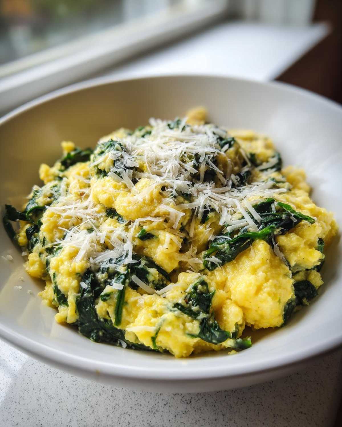 A close-up of One-Pot Creamy Spinach Parmesan Polenta topped with grated Parmesan cheese in a white bowl.