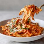 A fork lifting cheesy pieces of One-Pot Creamy Tomato Basil Tortilla Pasta from a white bowl.