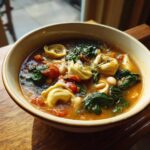 A close-up bowl of One-Pot Creamy Tortellini Minestrone soup featuring tortellini, spinach, tomatoes, and white beans.
