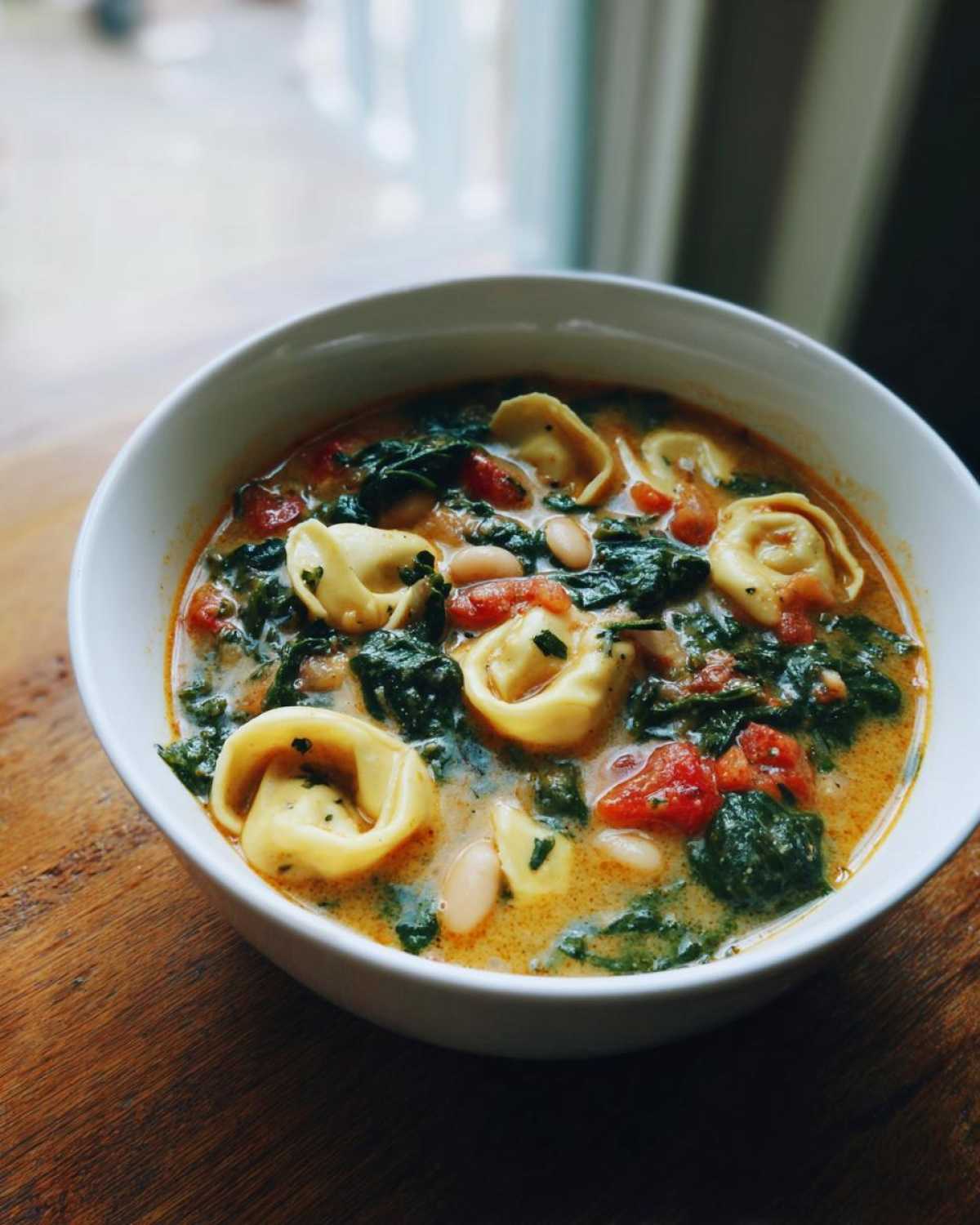 Close-up of a white bowl filled with One-Pot Creamy Tortellini Minestrone soup, featuring tortellini, spinach, tomatoes, and white beans.