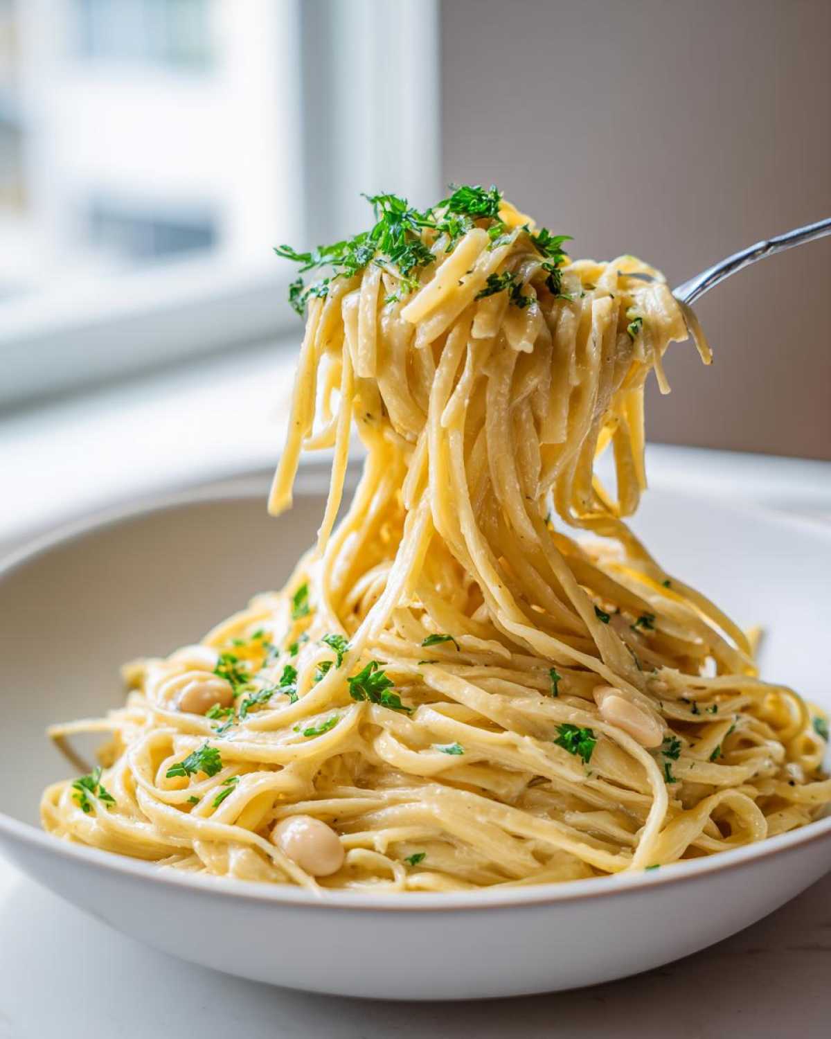 A fork lifts creamy strands of One-Pot Creamy White Bean Lemon Pasta, garnished with parsley, from a white bowl.