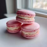 Three perfectly formed Pink Strawberry Macarons stacked and resting on a white plate near a window.