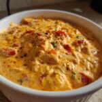 Close-up of a rich, orange Queso Dip in a white bowl, topped with peppers and spices.
