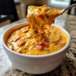 A spoonful of thick, cheesy Queso Dip, rich with peppers, is lifted from a white bowl.