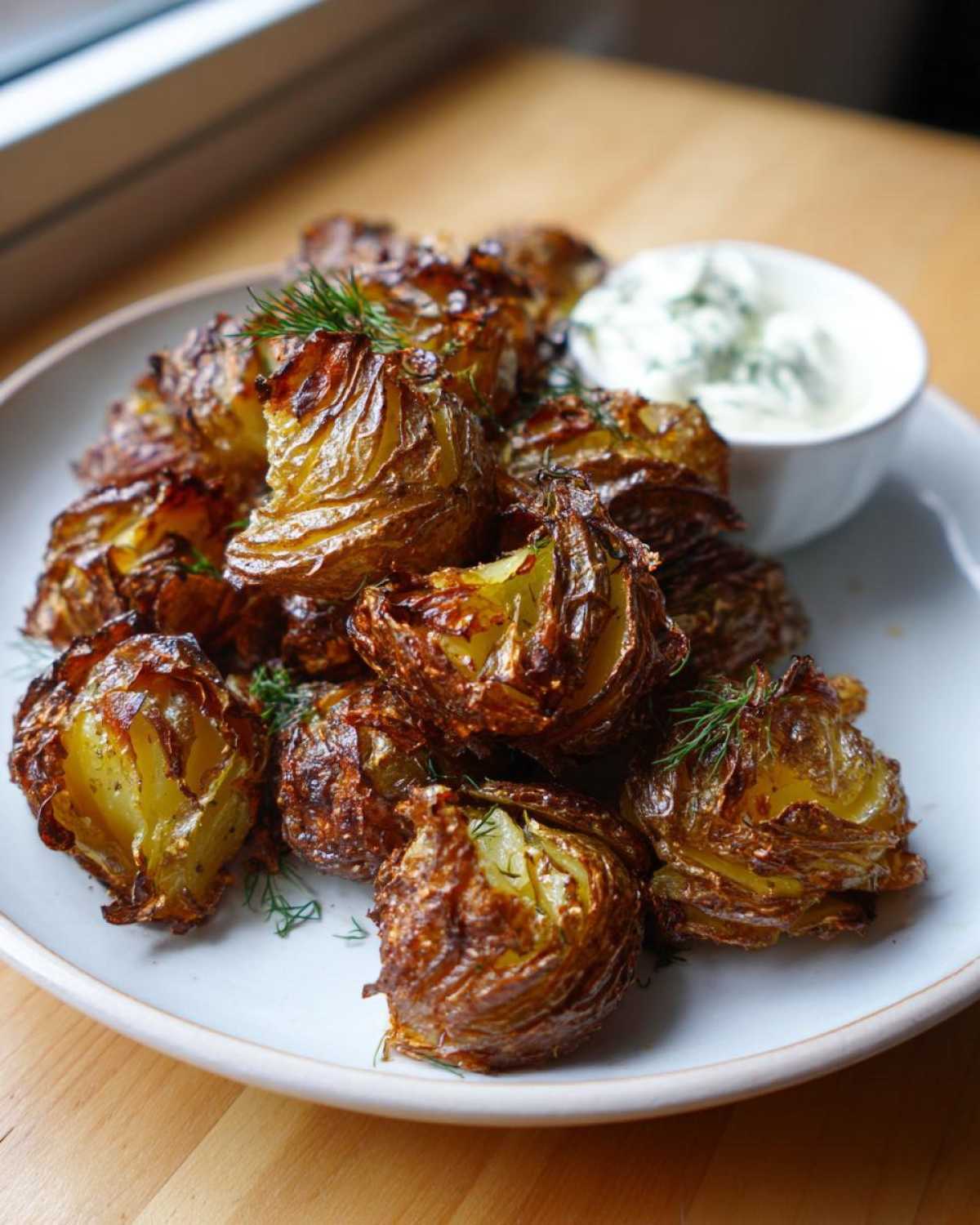 A plate of golden brown, crispy smashed potatoes garnished with dill, served alongside a small bowl of garlic aioli.